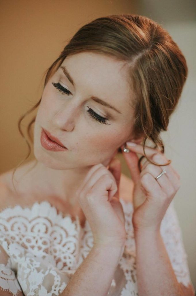 Bride with natural soft brown eyeshadow and nude lipstick putting in her earrings. Bridal makeup by Top Notch Art of Makeup.