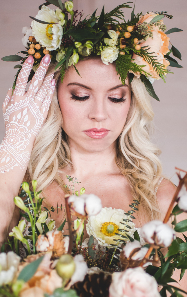 Bride with natural soft glam makeup looking down wearing a flower crown and touching her face with henna covered hands. Makeup by Top Notch Art of Makeup.