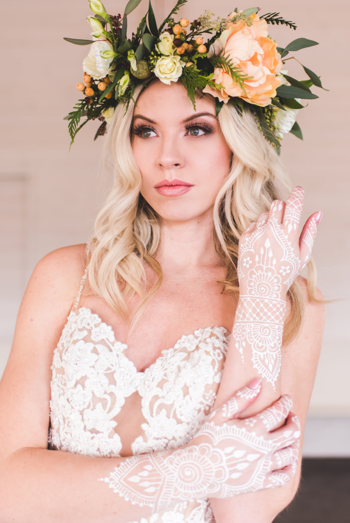 Bride wearing natural soft makeup with brown and pink tones looking into the distance. Her hand is touching her face with white henna she is wearing a yellow and green flower crown. Makeup by Top Notch Art of Makeup