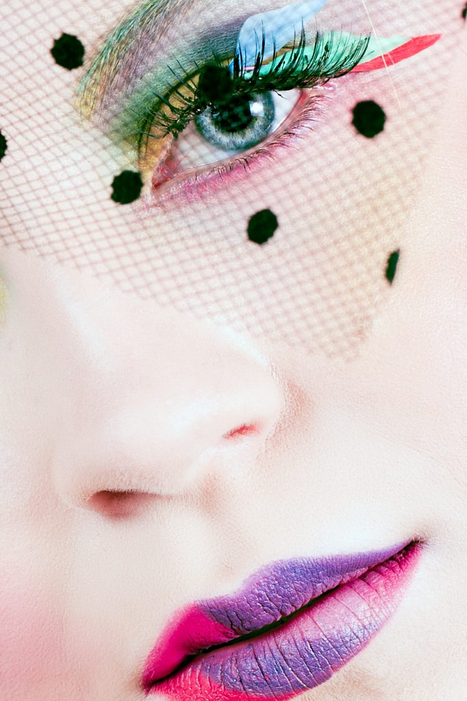 Close up of creative rainbow makeup on model with rainbow coloured feathers on eyelashes. A piece of polka dotted mesh is resting over the models eyes. Makeup by Top Notch Art of Makeup.