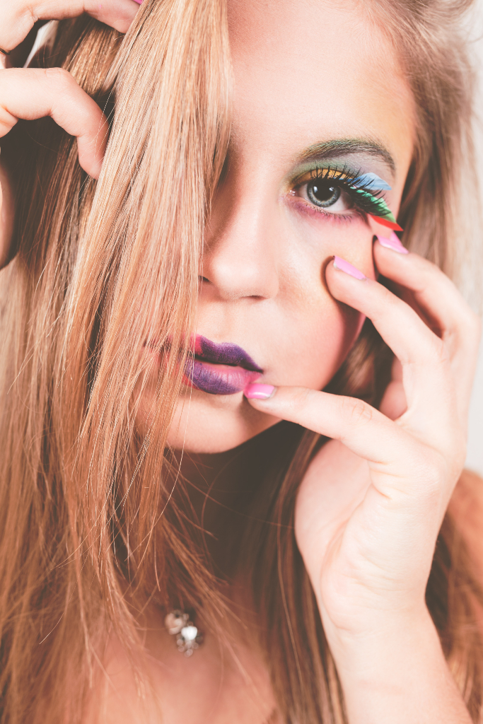 Close up portrait of model holding face with rainbow coloured eye shadows and rainbow feathers on eyes. Makeup by Top Notch Art of Makeup.