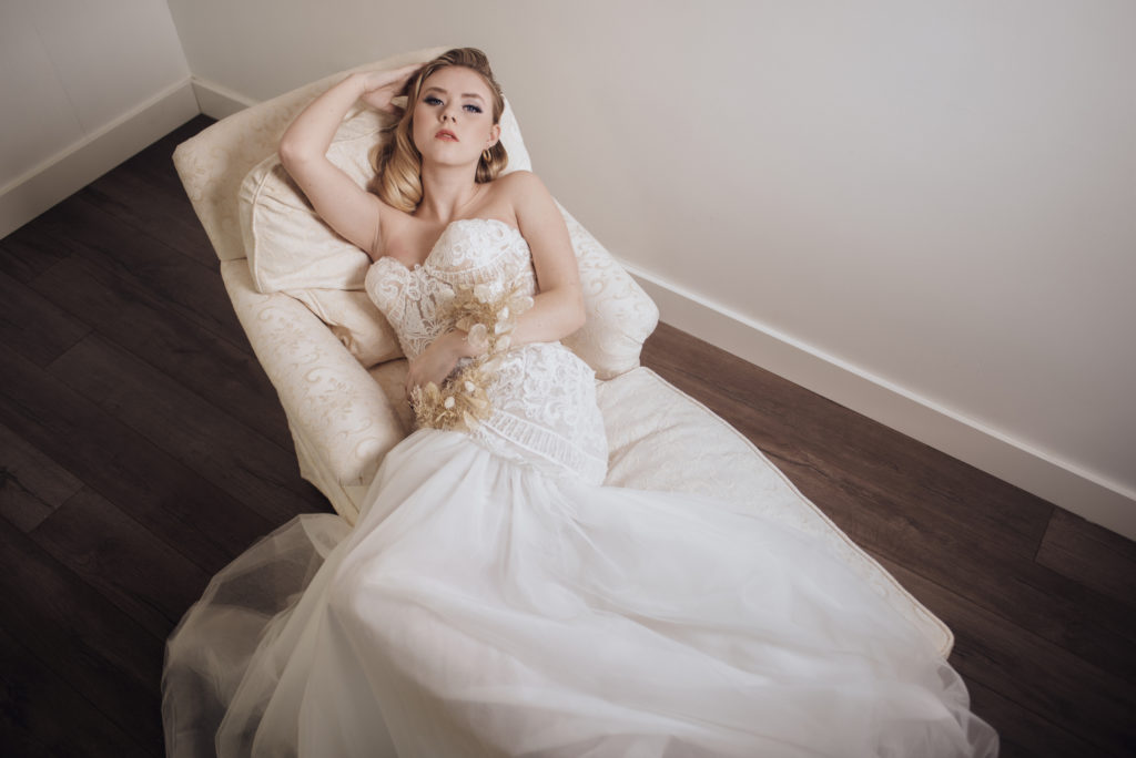 Bride in bridal gown laying down on a white couch with hand behind her head. Model is wearing natural makeup. Makeup by Top Notch Art of Makeup
