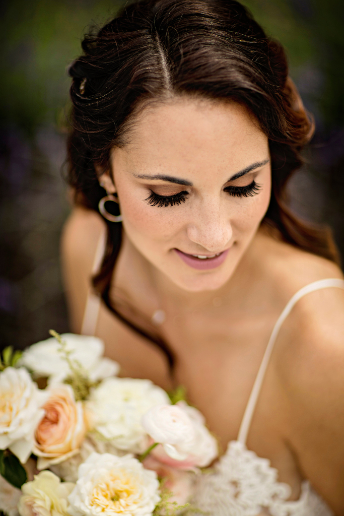 Bridal portrait of bride looking down with natural brown toned eye shadow and pink lipstick. Bride is holding a bouquet of white flowers. Bridal makeup by Top Notch Art of Makeup.