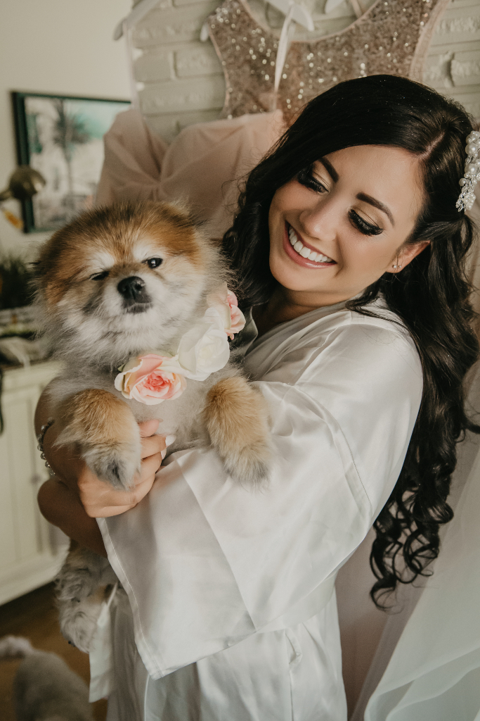 Bride with natural glam makeup holding and smiling at her puppy wearing a flower collar. Makeup by Top Notch Art of Makeup.