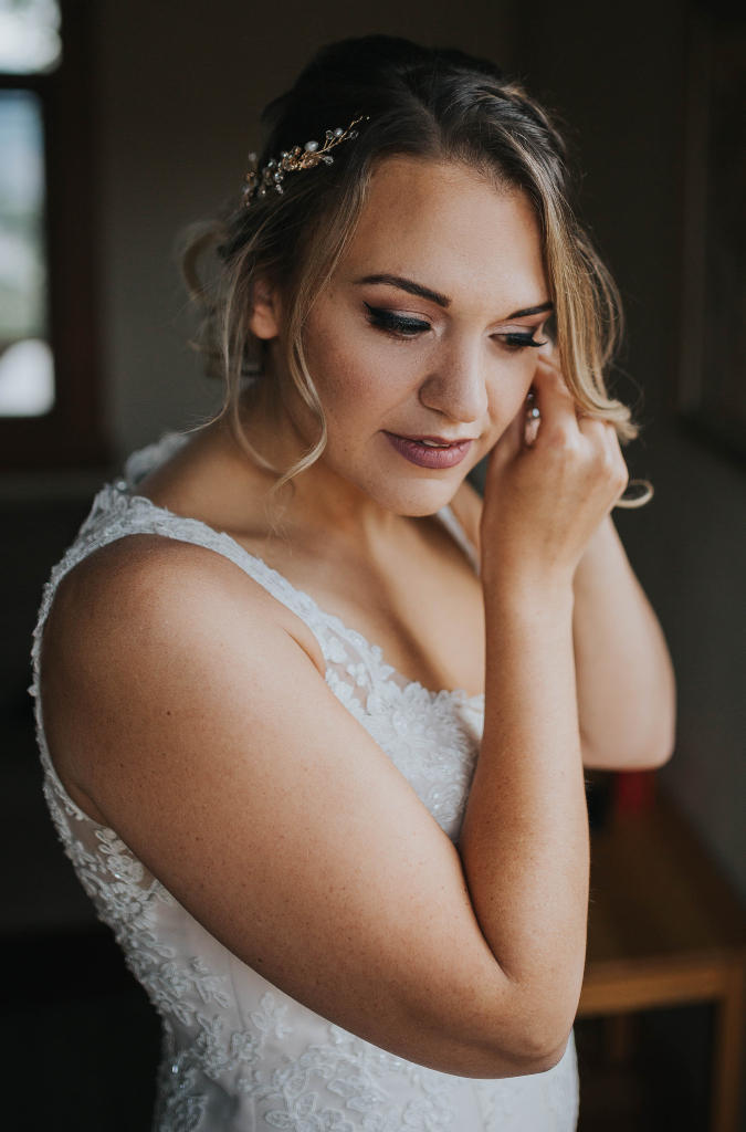 Portrait of Bride putting in earrings with pink toned makeup and floral hairpiece. Hair by Top Notch Art of Makeup