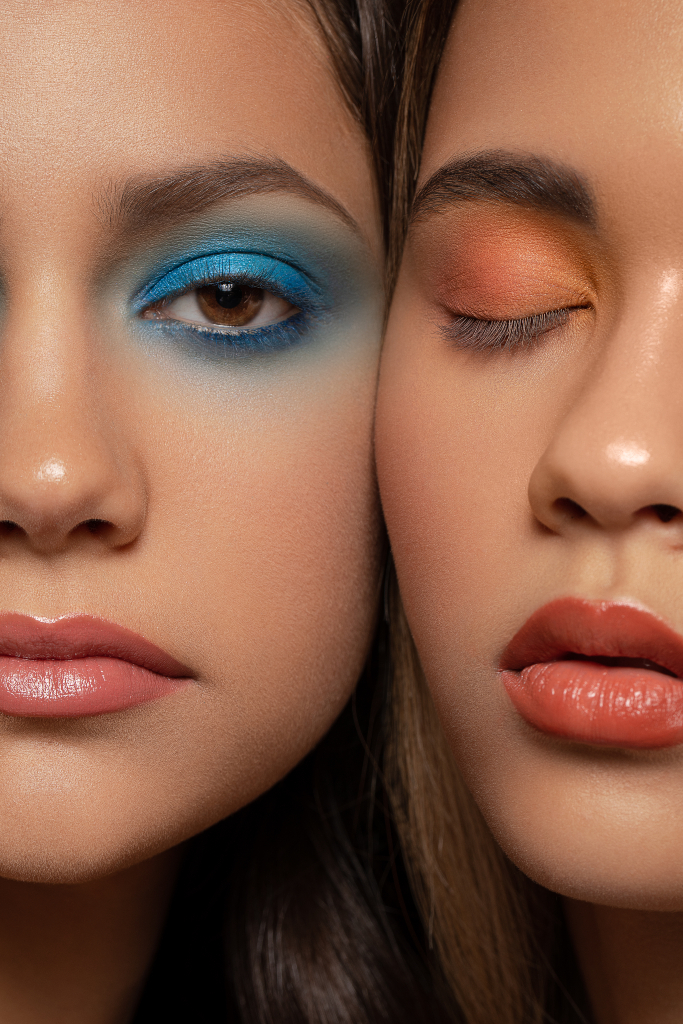 Close up portrait of two models faces, one models eye is open with blue eye makeup other model has eye closed with orange eyeshadow on eye. Makeup by Top Notch art of Makeup