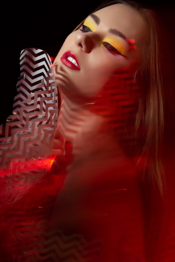 Creative yellow eyeshadow on model with winged eyeliner and red lipstick. Model is in red lighting and holding a piece of material with white and black lines on it. Makeup by Top Notch Art of Makeup.