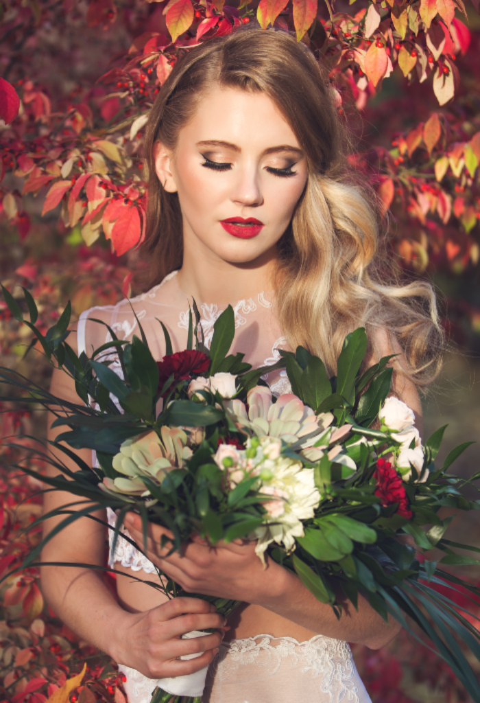 Bride wearing glam makeup with a brown smokey eye and bold red lips. Bride is holding a bouquet with pops of red flowers in a forest with red toned trees. Makeup by Top Notch Art of Makeup