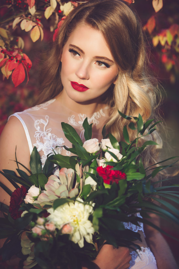 Bride wearing glam classic makeup with winged eyeliner and bold red lip. Bride is in the forest with red coloured trees holding a bouquet of flowers. Makeup by Top Notch Art of Makeup.