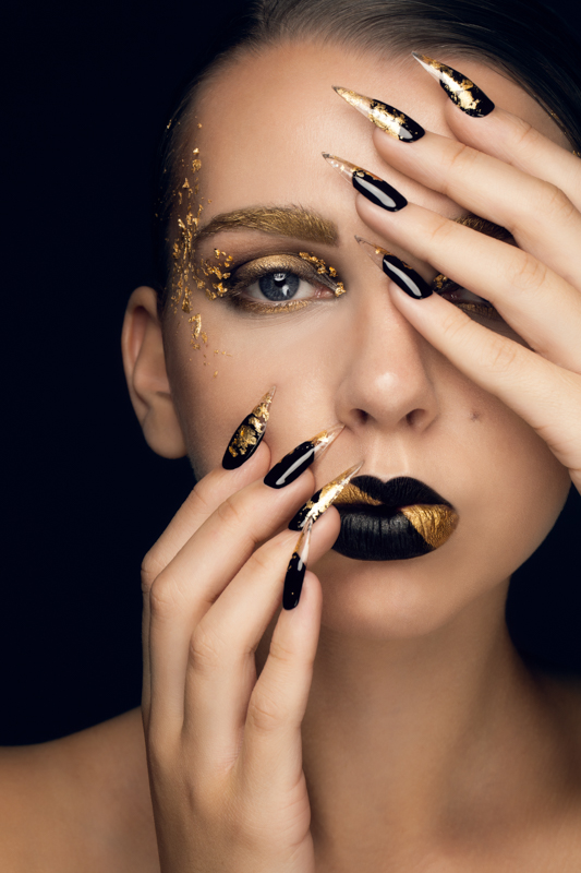 Creative black and gold makeup on model with gold flakes surrounding eyes. Both of models hands are on her face with gold and black long nails. Makeup by Top Notch Art of Makeup